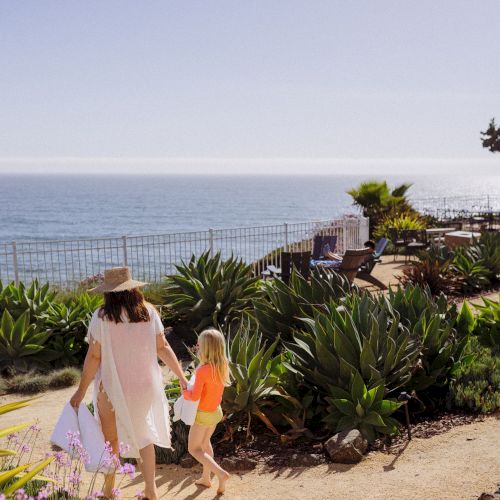 A woman and child stroll in a seaside garden with benches and lounge chairs, looking out over the ocean.