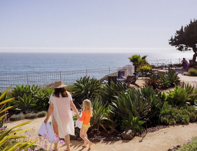 A woman and child stroll in a seaside garden with benches and lounge chairs, looking out over the ocean.